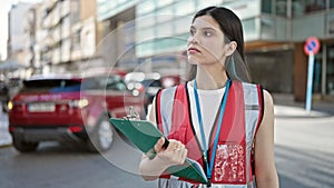 Young beautiful hispanic woman survey interviewer holding clipboard standing with serious expression at street