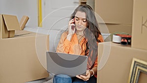 Young beautiful hispanic woman talking on smartphone using laptop at new home