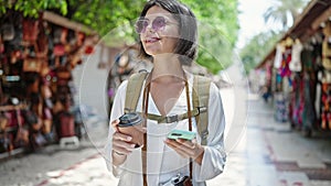 Young beautiful hispanic woman tourist smiling confident using smartphone drinking coffee at street market