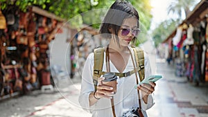 Young beautiful hispanic woman tourist using smartphone drinking coffee at street market