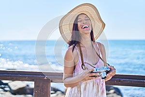 Young beautiful hispanic woman tourist using vintage camera at seaside
