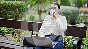 Young beautiful hispanic woman using laptop sitting on a bench drinking coffee at the park