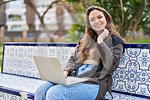 Young beautiful hispanic woman using laptop sitting on bench at park