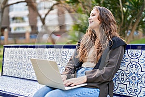 Young beautiful hispanic woman using laptop sitting on bench at park