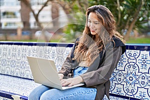 Young beautiful hispanic woman using laptop sitting on bench at park