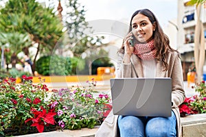 Young beautiful hispanic woman using laptop talking on smartphone at park