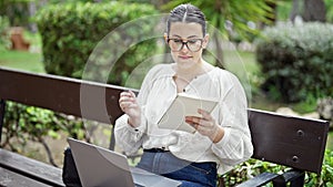 Young beautiful hispanic woman using laptop writing on a notebook sitting on a bench at the park