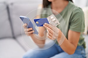 Young beautiful hispanic woman using smartphone and credit card holding pills at home