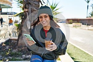 Young beautiful hispanic woman using smartphone drinking coffee at park