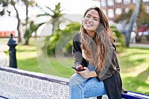 Young beautiful hispanic woman using smartphone sitting on bench at park