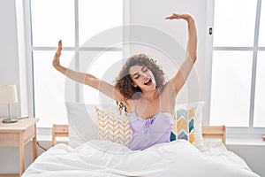Young beautiful hispanic woman waking up stretching arms yawning at bedroom