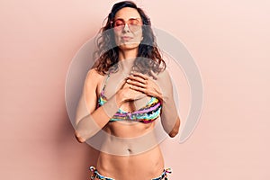 Young beautiful hispanic woman wearing bikini and heart sunglasses smiling with hands on chest, eyes closed with grateful gesture