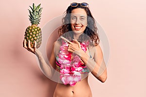 Young beautiful hispanic woman wearing bikini holding pineapple smiling happy pointing with hand and finger