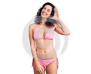 Young beautiful hispanic woman wearing bikini waiving saying hello happy and smiling, friendly welcome gesture