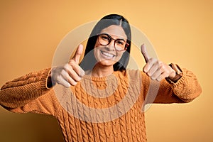 Young beautiful hispanic woman wearing glasses over yellow isolated background approving doing positive gesture with hand, thumbs