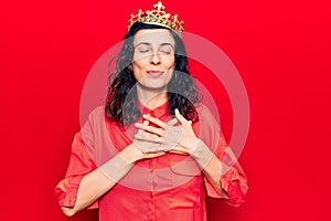 Young beautiful hispanic woman wearing princess crown smiling with hands on chest, eyes closed with grateful gesture on face