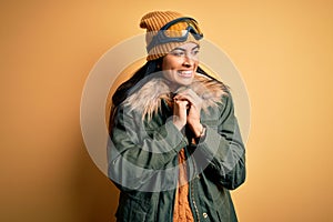 Young beautiful hispanic woman wearing ski glasses and coat for winter weather laughing nervous and excited with hands on chin