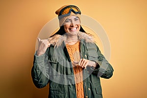 Young beautiful hispanic woman wearing ski glasses and coat for winter weather Pointing to the back behind with hand and thumbs