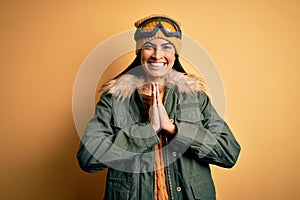 Young beautiful hispanic woman wearing ski glasses and coat for winter weather praying with hands together asking for forgiveness