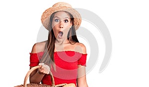 Young beautiful hispanic woman wearing summer hat and holding picnic wicker basket with bread scared and amazed with open mouth