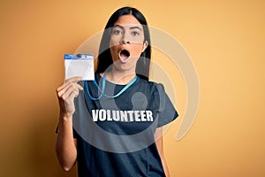 Young beautiful hispanic woman wearing volunteer t-shirt and showing identification scared in shock with a surprise face, afraid