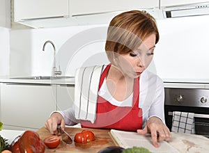 Young beautiful home cook woman in red apron at domestic kitchen reading cookbook following recipe holding knife