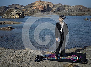 Young beautiful indian girl reading a book sitting on a blanket in the mountains with a lake nearby. Concept of having picnic in a