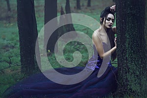 Young beautiful lady in luxurious sequin evening dress sitting in the mysterious misty woods leaning on the moss covered tree