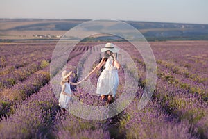 Young beautiful lady mother with lovely daughter walking on the lavender field on a weekend day in wonderful dresses and hats.