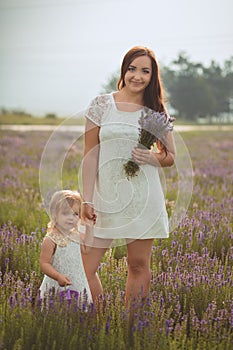 Young beautiful lady mother with lovely daughter walking on the lavender field on a weekend day in wonderful dresses and hats.