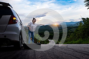 Young beautiful lady standing near car for calling for help on the public road in forest area at mountain and sky