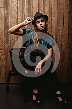 Young beautiful lady in a stylish hat and casual clothes holds a hat with her hand, sits in a chair and looks at camera