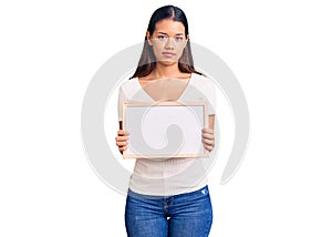 Young beautiful latin girl holding empty white chalkboard thinking attitude and sober expression looking self confident