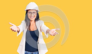 Young beautiful latin girl wearing architect hardhat and glasses looking at the camera smiling with open arms for hug