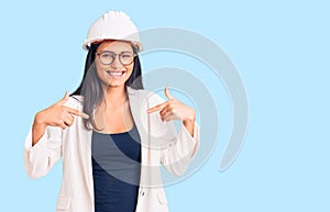 Young beautiful latin girl wearing architect hardhat and glasses looking confident with smile on face, pointing oneself with