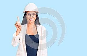 Young beautiful latin girl wearing architect hardhat and glasses showing and pointing up with finger number one while smiling