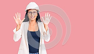 Young beautiful latin girl wearing architect hardhat and glasses showing and pointing up with fingers number ten while smiling