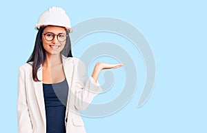 Young beautiful latin girl wearing architect hardhat and glasses smiling cheerful presenting and pointing with palm of hand