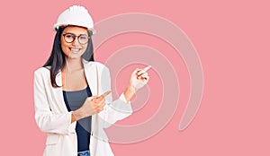 Young beautiful latin girl wearing architect hardhat and glasses smiling and looking at the camera pointing with two hands and