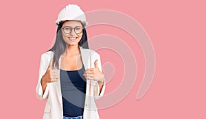 Young beautiful latin girl wearing architect hardhat and glasses success sign doing positive gesture with hand, thumbs up smiling