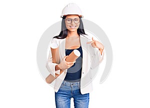 Young beautiful latin girl wearing architect hardhat holding blueprints pointing finger to one self smiling happy and proud