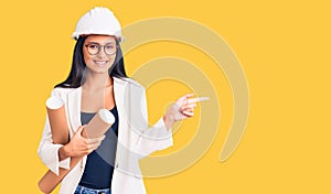 Young beautiful latin girl wearing architect hardhat holding blueprints smiling happy pointing with hand and finger to the side