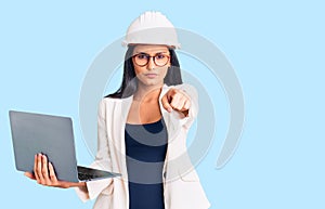 Young beautiful latin girl wearing architect hardhat holding laptop pointing with finger to the camera and to you, confident