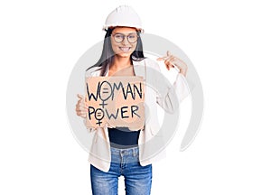 Young beautiful latin girl wearing architect hardhat holding woman power banner pointing finger to one self smiling happy and