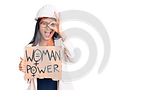 Young beautiful latin girl wearing architect hardhat holding woman power banner smiling happy doing ok sign with hand on eye