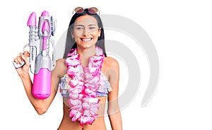 Young beautiful latin girl wearing bikini and hawaiian lei holding water gun looking positive and happy standing and smiling with