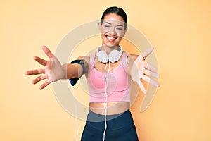 Young beautiful latin girl wearing gym clothes and using headphones looking at the camera smiling with open arms for hug