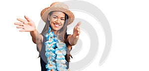 Young beautiful latin girl wearing hawaiian lei and summer hat looking at the camera smiling with open arms for hug