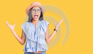 Young beautiful latin girl wearing summer hat and glasses crazy and mad shouting and yelling with aggressive expression and arms