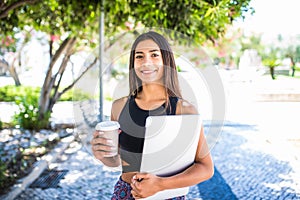 Young beautiful latin student with laptop and cup of coffee studying in the park. Girl is walking in the park with great smile and